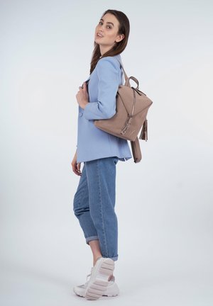 Beige Stoff-Rucksack mit dreieckigem Design und silbernem Zubehör, mit einem Reißverschluss und Quastenakzenten, lässig getragen mit einem blauen Blazer und Jeans.