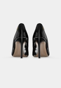 Chaussures à talons hauts en cuir verni noir vues de dos, avec de hauts talons aiguilles minces sur un fond clair uni.