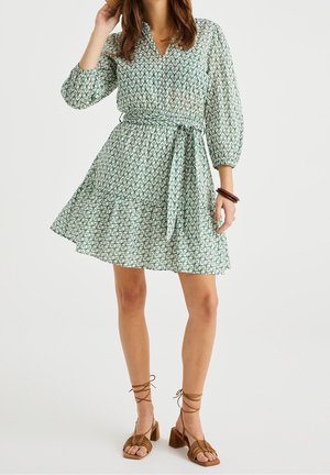 Groen jurk met patroon, V-hals, lange pofmouwen en een taille met ceintuur. Uitlopende rok met een gerimpelde zoom. Gecombineerd met bruine sandalen met riempjes.