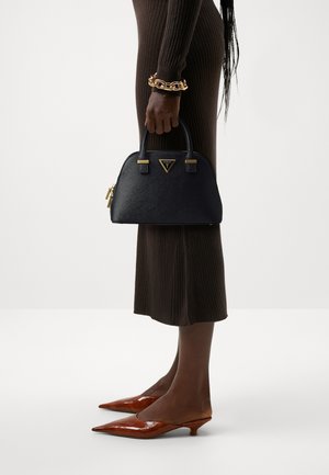 Une personne portant une robe en maille marron côtelée et un bracelet doré tient un sac à main noir Guess, portant des chaussures marron pointues à petit talon sur fond blanc.