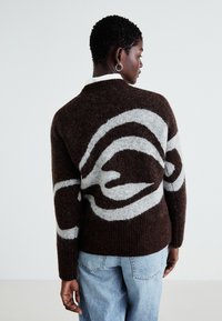 Brun strikkegenser med et hvitt swirl-mønster. Har ribbestrikket søm og mansjetter, samt rund hals, kombinert med lyseblå jeans.