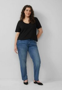Schwarzes T-Shirt mit kurzen Ärmeln und Rundhalsausschnitt, kombiniert mit blauen geraden Jeans und schwarzen Ballerinas. Das Model steht vor einem neutralen Hintergrund.