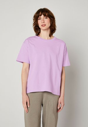 Lila T-Shirt mit kurzen Ärmeln in entspannter Passform, aus glattem Baumwollmaterial mit Rundhalsausschnitt, kombiniert mit hellbeigen, weit geschnittenen Hosen.
