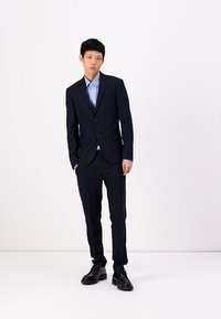 Navy pinstripe suit with two buttons, worn over a light blue shirt. Features a tailored fit and paired with black dress shoes.