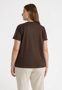 Camiseta corta de manga, color marrón, hecha de algodón suave, con un corte relajado y un escote redondo, combinada con pantalones de color claro.