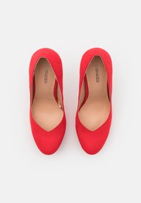Escarpins en cuir suédé rouge avec un design à bout pointu, présentant une découpe sur le côté et une texture intérieure lisse. Grand logo de la marque visible sur la semelle intérieure.