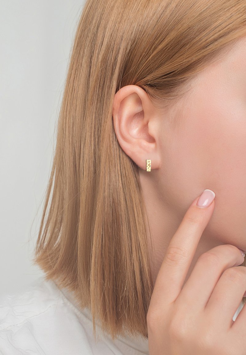 Orecchino a cerchio in oro con superficie testurizzata, indossato all'orecchio di un modello. Capelli biondi corti e lisci e un primo piano di una mano che tocca il viso.