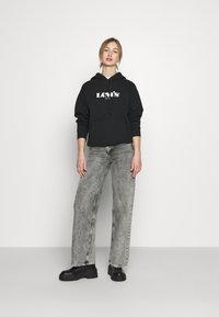 Felpa nera con logo bianco "Levi's", abbinata a jeans grigi chiari a gamba larga. L'outfit presenta una vestibilità rilassata e colori solidi. Calzature nere completano il look.