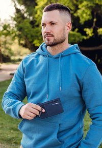 Navy leather wallet held by a person, featuring a card slot and key attachment, with a blue hoodie in a green park setting.
