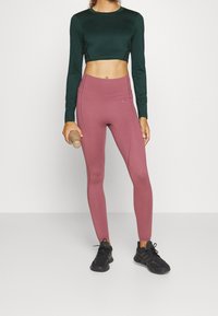 Vihreä pitkähihaine crop-top, yhdistettynä korkeavyötäröisiin vaaleanpunaiseen leggingsiin. Malli pitää kädessään vesipulloa ja hänellä on jalassaan mustat urheilukengät.