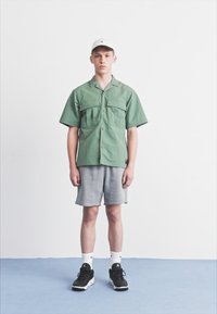 Grön kortärmad skjorta med knappar och två bröstfickor, i kombination med grå shorts och svarta sneakers. Vit keps bärs på huvudet.