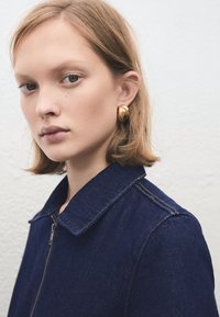 Džínsová bunda tmavomodrej farby so zapínaním na zips, špicatým golierom a kontrastným prešitím. Modelka má veľké zlaté kruhové náušnice.