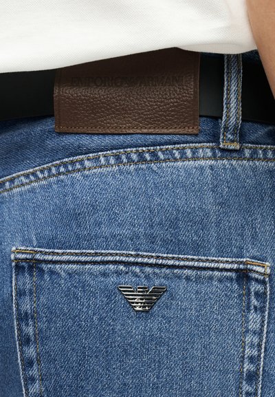 Κοντινό πλάνο τσέπης μπλε τζιν με μεταλλικό λογότυπο Emporio Armani και καφέ δερμάτινο ταμπελάκι επωνυμίας στη ζώνη μέσης.