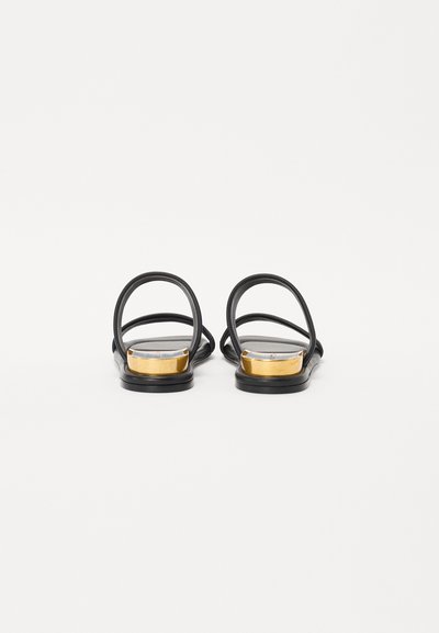 Sandalias planas negras con diseño de doble tira, acento dorado en el talón y material suave, con una estructura minimalista y abierta por detrás.