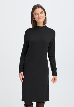 Femme aux cheveux châtain mi-longs portant une robe noire à manches longues, des collants noirs et des bottes marron, debout devant un fond uni.