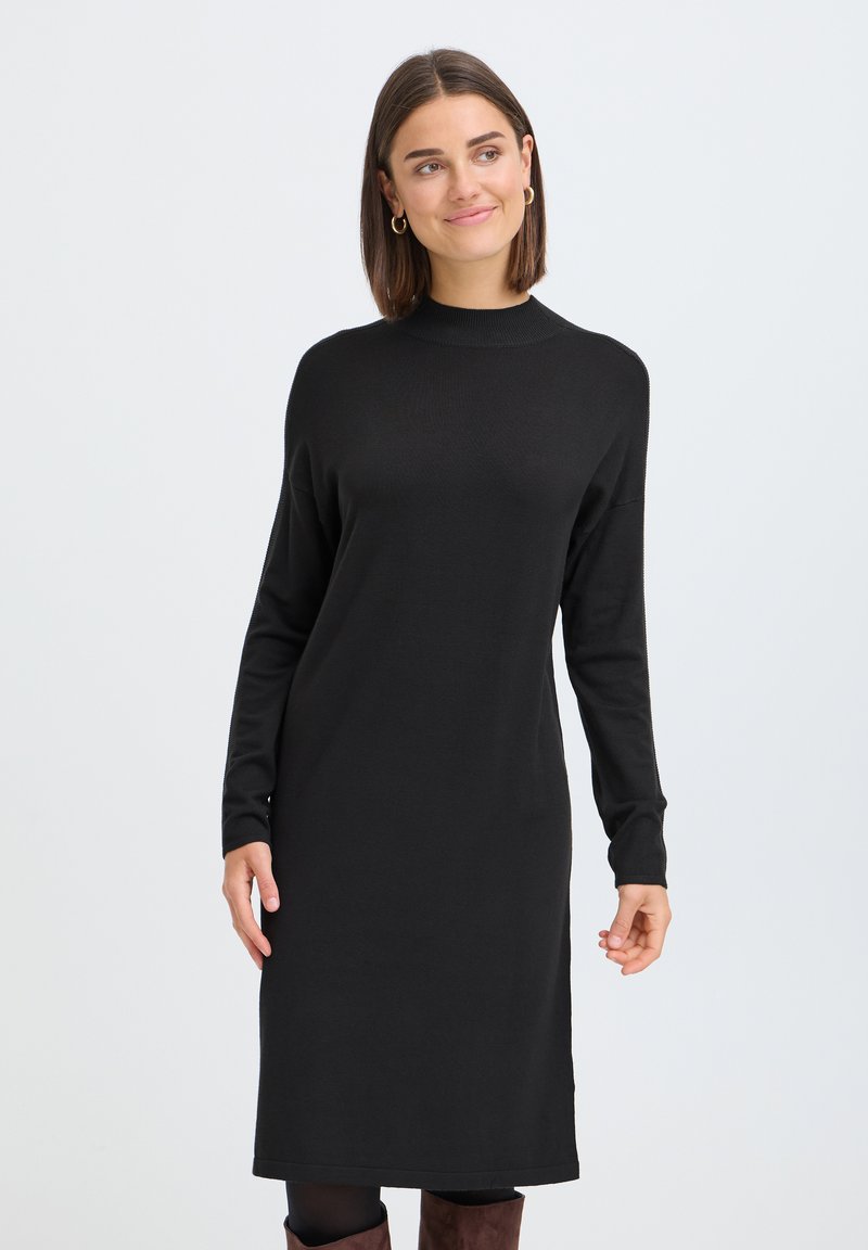 Femme aux cheveux châtain mi-longs portant une robe noire à manches longues, des collants noirs et des bottes marron, debout devant un fond uni.