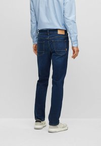 Dark blue denim jeans with a tapered fit, featuring a brown leather logo patch, back pockets, and contrast stitching.