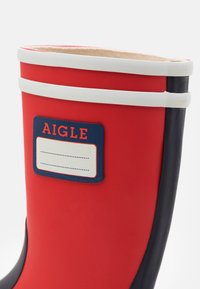 Red rubber boot with a smooth texture, designed with white and navy accents, featuring a rectangular label displaying the brand name "Aigle."
