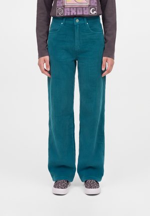 Personne portant un pantalon large en velours côtelé bleu sarcelle, un t-shirt graphique à manches longues foncé et des chaussures blanches à enfiler avec imprimé léopard noir.