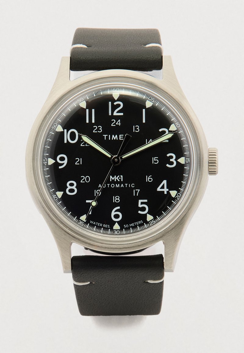 Montre analogique avec cadran noir, chiffres blancs (12 heures et 24 heures), aiguilles luminescentes, boîtier en acier inoxydable et bracelet en cuir noir.