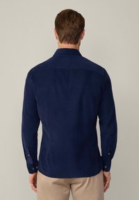 Hombre con cabello corto castaño, vistiendo una camisa de manga larga azul marino y pantalones beige, mostrado de espaldas contra un fondo liso.