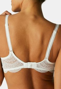Weißer Spitzentop mit verstellbaren Trägern und einem dekorativen Rückenband. Verfügt über florale Muster und gewellte Kanten. Weiche Stofftextur.