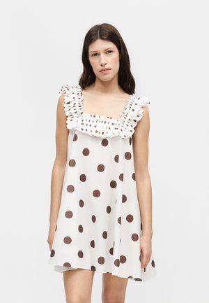 Kobieta w białej sukience bez rękawów z dużymi brązowymi groszkami i marszczonym kwadratowym dekoltem, stojąca na jednolitym jasnym tle.