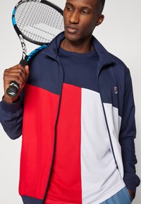 Men's athletic outfit with a navy jacket featuring red and white panels, paired with a navy top, holding a tennis racket.