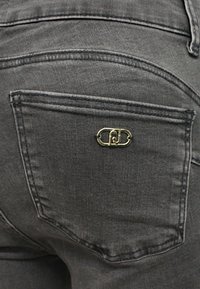 Jeans in denim grigio scuro con un distintivo logo dorato sulla tasca posteriore, caratterizzati da cuciture standard e una texture liscia.