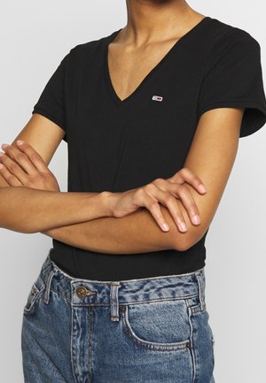 Personne portant un t-shirt noir à col en V avec un petit logo sur la poitrine et un jean bleu, bras croisés.
