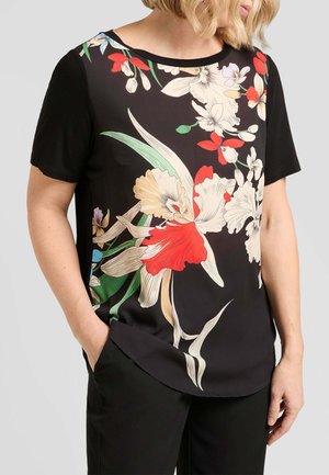 Donna che indossa una maglia nera a maniche corte con una grande stampa floreale rossa, bianca, verde e beige, abbinata a pantaloni neri.