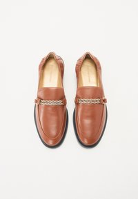 Brūni ādas loafers, kas izceļas ar pītas zelta tonis ķēdes akcentu pāri pirkstiem, gluda virsma, noapaļots pirksts un melns gumijas zole.