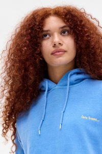 Junge Frau mit langen lockigen roten Haaren, die einen blauen Hoodie mit gelbem Text "lets frans..." trägt und leicht nach oben schaut.