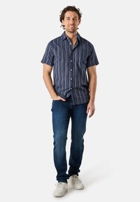 Korte mouwen marineblauw shirt met verticale strepen, knoopsluiting aan de voorkant, borstzak, gedragen met donkerblauwe spijkerbroek en witte sneakers.