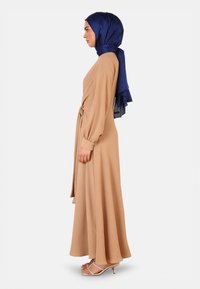 Beige long dress with long sleeves and a tied waist, made of textured material. Paired with a dark blue satin hijab and nude heels.