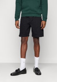 Pantalones cortos de algodón negros con bolsillos laterales, combinados con una sudadera verde, y acompañados de unos mocasines negros y calcetines blancos con un pequeño logo.