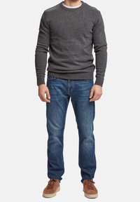 Homme portant un pull gris foncé ajusté, un jean bleu et des baskets en daim marron, sur un fond blanc uni.