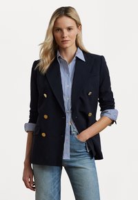 Marineblauw double-breasted blazer met gouden knopen, gedragen over een blauw-wit gestreept shirt en lichtblauwe jeans. Glad materiaal, perfecte pasvorm.