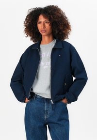 Donkerblauwe bomberjack met ritssluiting aan de voorkant, elastische manchetten en zijzakken, gelaagd over een grijze shirt met paarse letters. Denim jeans aanwezig.