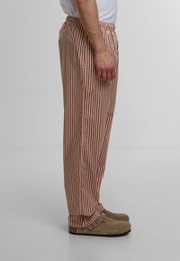 Pantaloni rayati cu un design roșu și bej, bretele elastice și croială lejeră, asortați cu saboți bej. Textura pare netedă și ușoară.