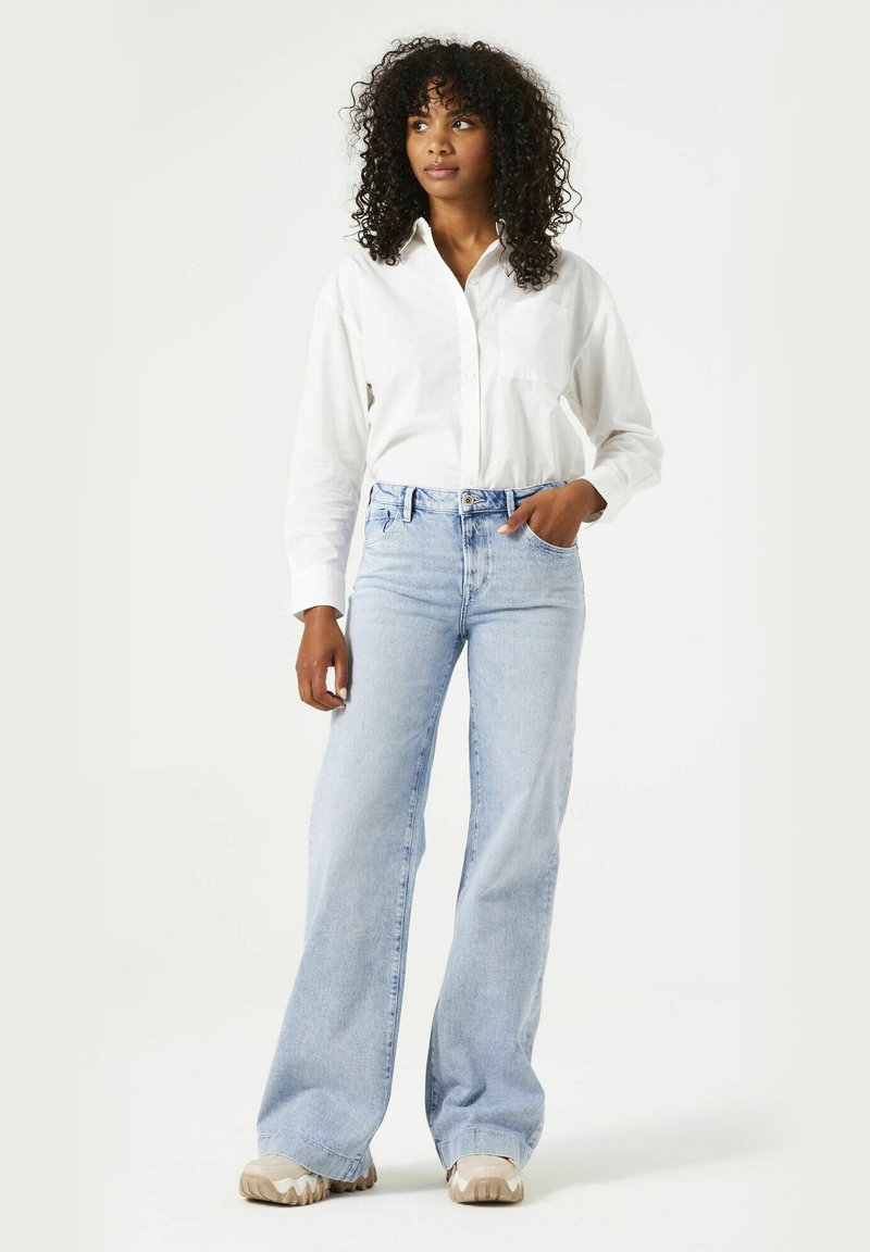 Jeans de tiro alto en azul claro con un corte relajado, combinados con una camisa blanca de botones. El conjunto presenta un diseño minimalista y una textura suave.