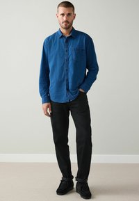 Blå skjorta med knappknäppning, texturerad yta och bröstficka, kombinerad med svarta byxor och svarta skor, stående mot en neutral bakgrund.