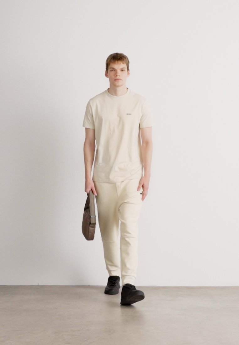 T-shirt beige clair et joggings assortis, en tissu doux. La tenue est accompagnée de chaussures noires et d'un sac à bandoulière marron.