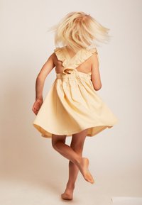 Helles gelbes Kleid mit ausgestellt Rock, Rüschenträgern und gerafftem Oberteil, von hinten sichtbar, mit baren Füßen.