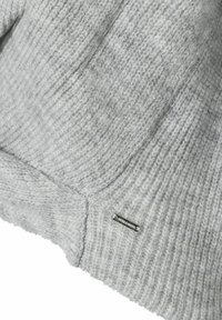 Tissu en maille gris clair avec texture côtelée et petite étiquette métallique rectangulaire fixée près du bord inférieur.