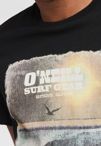 Camiseta de algodón negra con un gráfico de estilo vintage que incluye el texto "O'Neill Surf Gear", imágenes de paisajes y un acento de un pájaro volando.