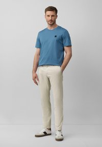 Niebieska koszulka z krótkim rękawem z kwadratowym detalem logo, sparowana z jasnobeżowymi spodniami i białymi sneakerami z czarnymi akcentami.