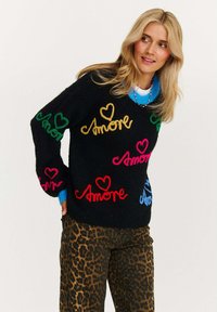 Czarny sweter z kolorowym napisem "Amore" oraz motywami serc w kolorach różowym, zielonym, żółtym i niebieskim; posiada szerokie rękawy i luźny krój. Połączony z spodniami w panterkę.