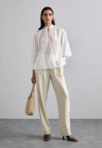 Blouse blanche légère avec encolure froncée et manches longues, associée à un pantalon large crème et des escarpins pointus marron. Sac à épaule beige.