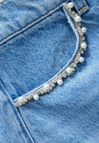 Buzunar din denim bleu deschis cu margine decorativă, având perle false și pietre prețioase transparente. Detalii vizibile de cusătură de-a lungul marginilor buzunarului.